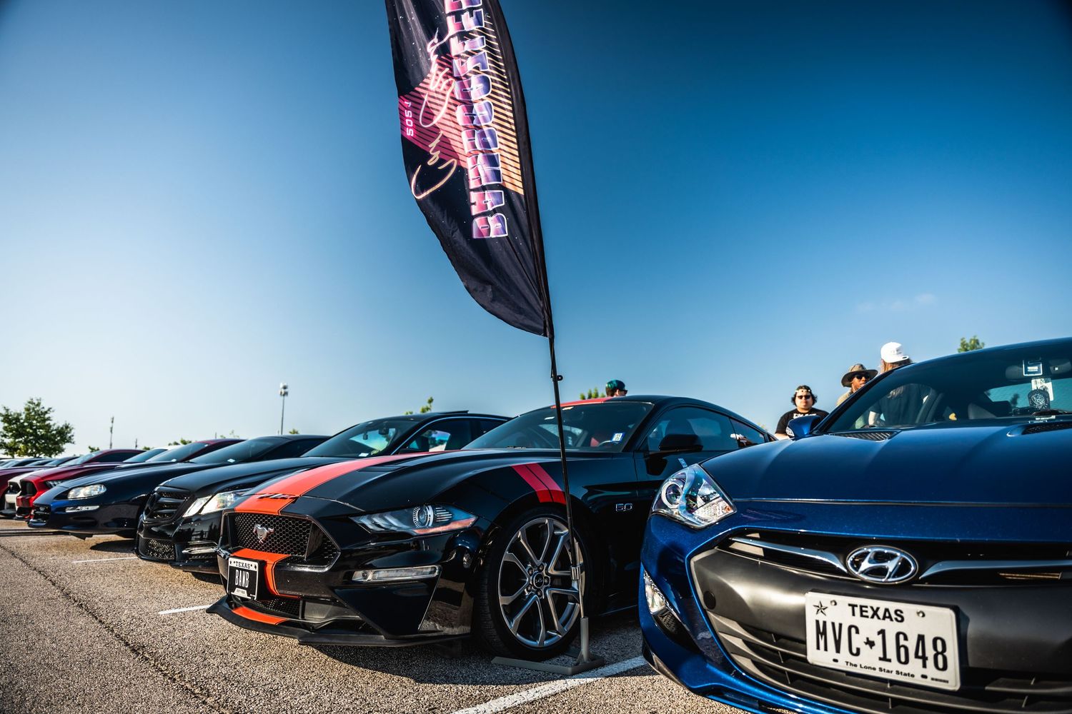 Line of performance cars including Mustangs and Hyundai vehicles at an outdoor car meet.