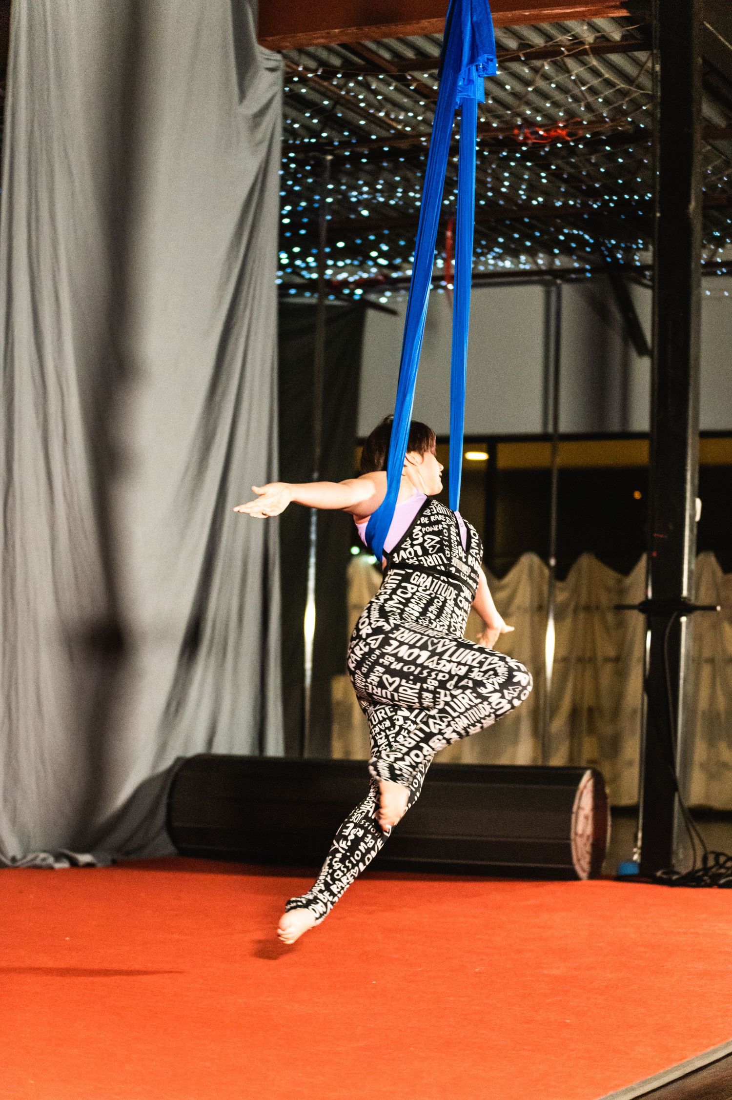 Performer in patterned outfit executes dynamic pose on blue aerial silks against dark background.