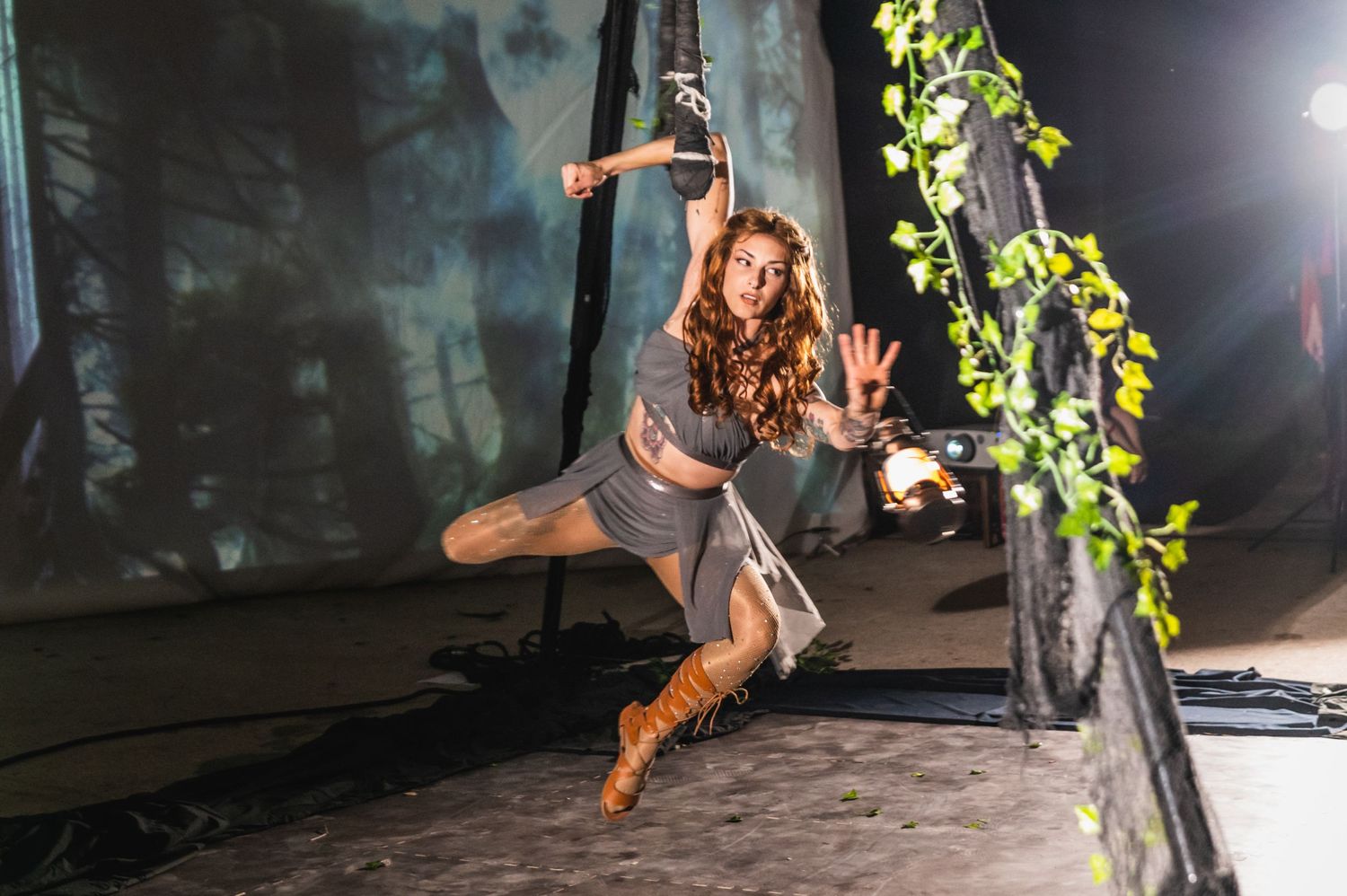 Aerial artist in gray costume performs split pose on suspended equipment under stage lighting.