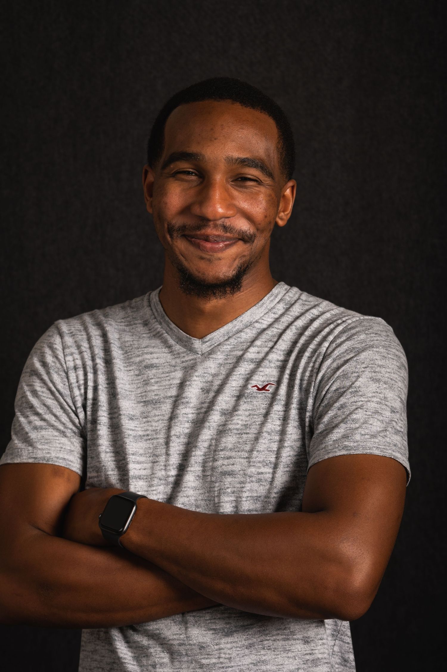 Professional headshot of a person in a gray v-neck t-shirt smiling warmly against a dark background.