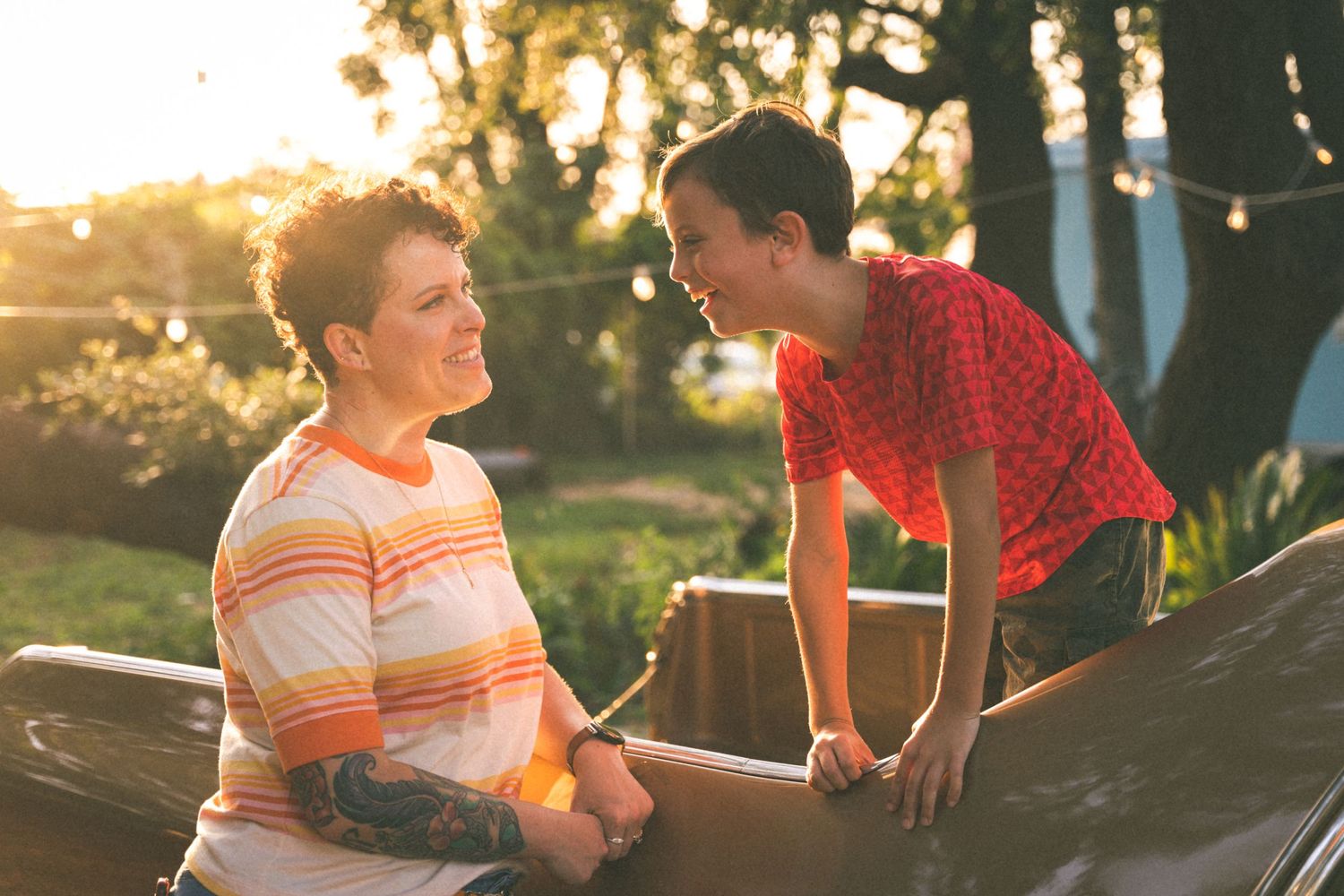 Two figures conversing outdoors at sunset with golden sunlight filtering through trees in the background.
