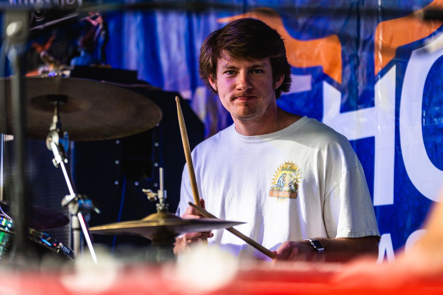 Drummer performs behind drum kit on stage with blue background decoration.