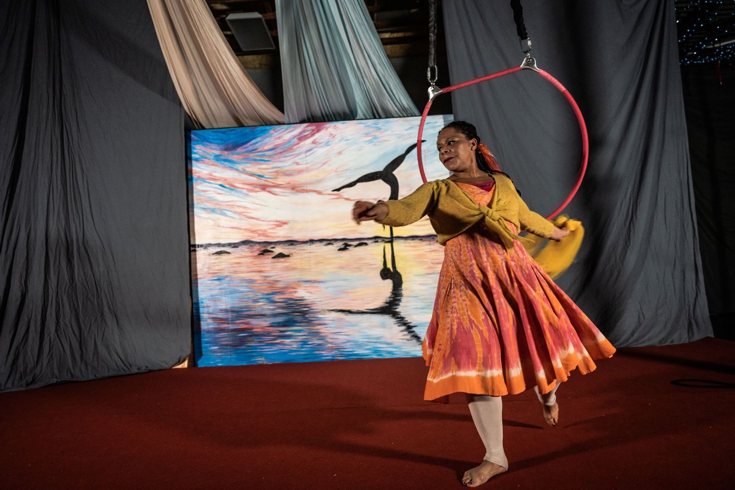 Performer in red dress dances with ribbon against painted sunset backdrop.