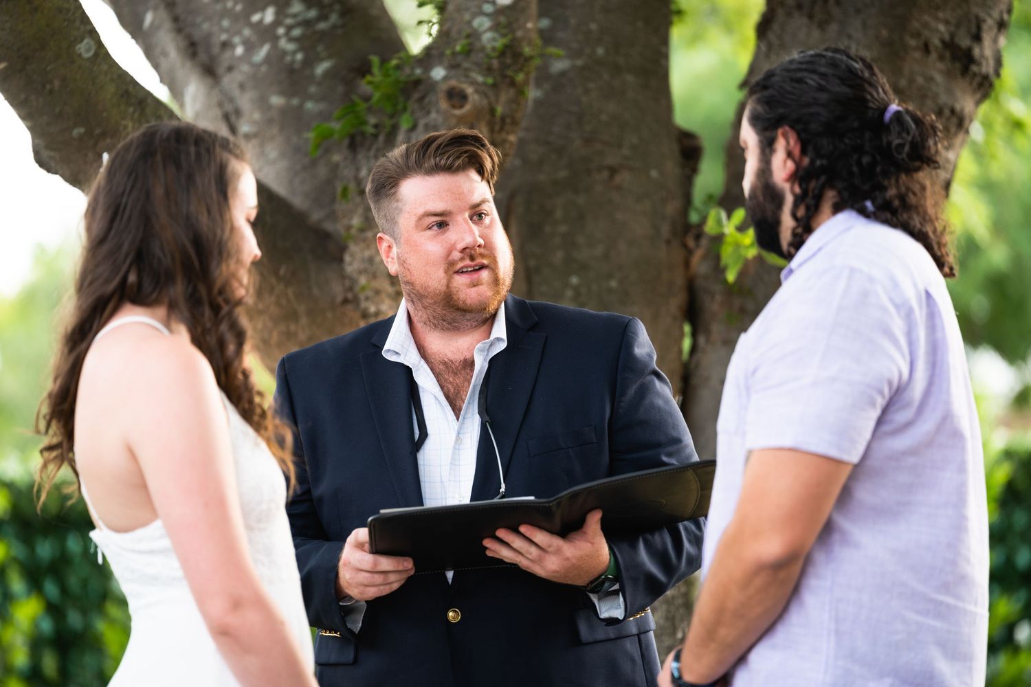 An outdoor wedding ceremony takes place under trees with an officiant speaking to a couple.