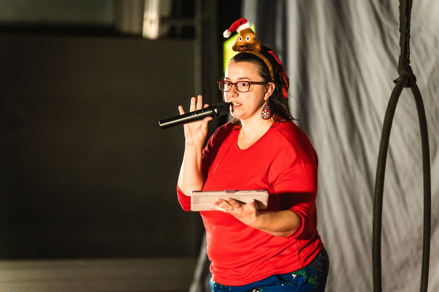 Performer in red shirt and festive hat singing into microphone on dimly lit stage.