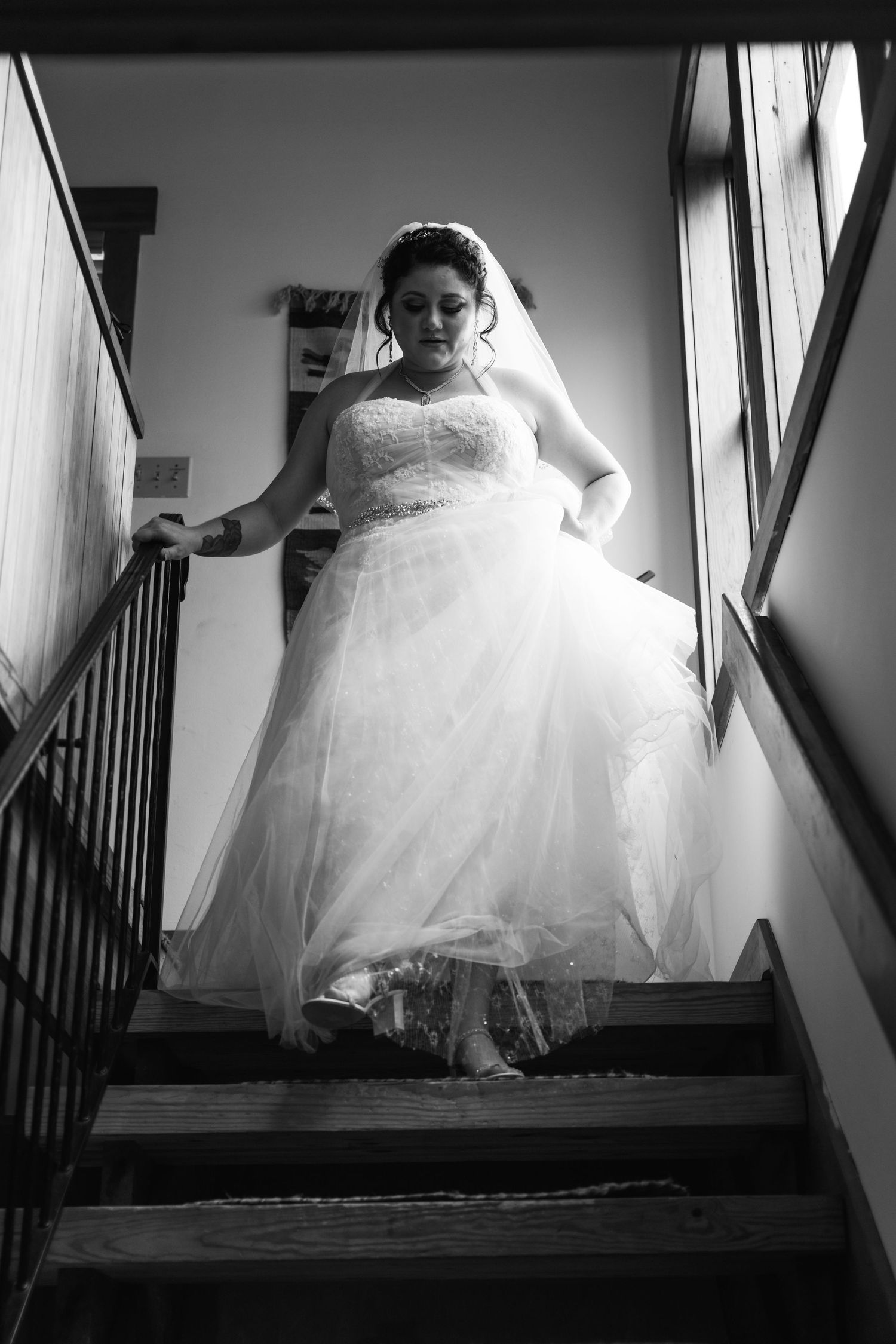 A bride in a lace wedding dress descends a staircase in dramatic black and white lighting.