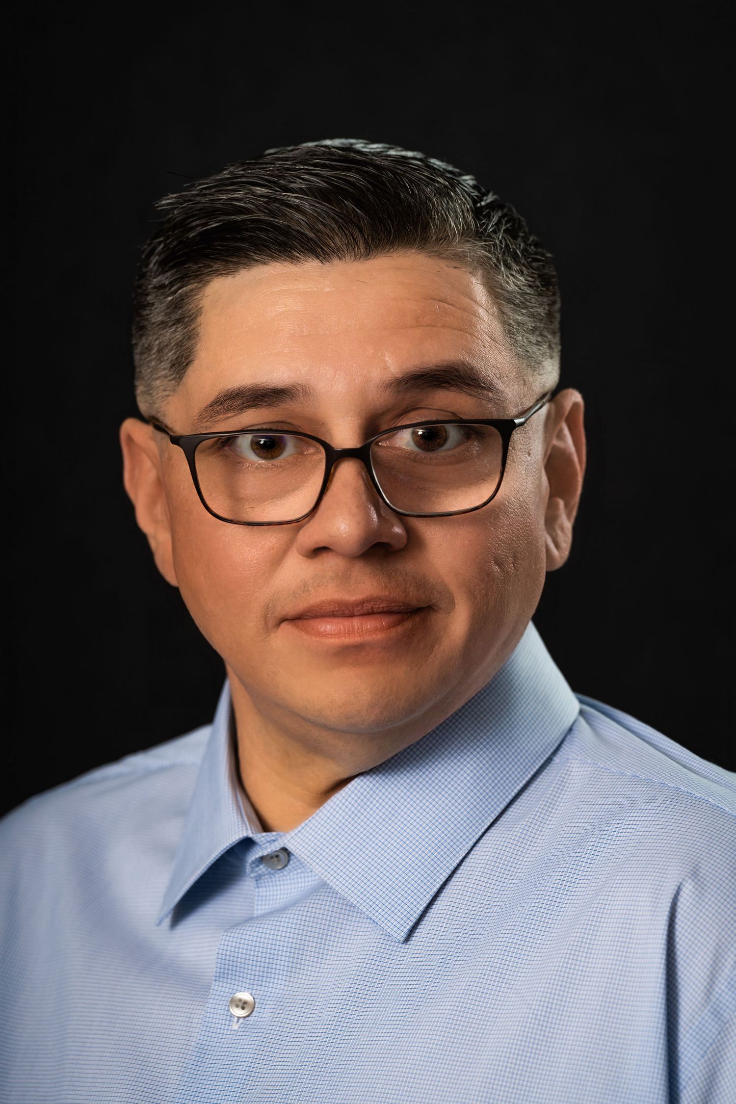 Professional headshot of a person wearing glasses and a light blue collared shirt against dark background.