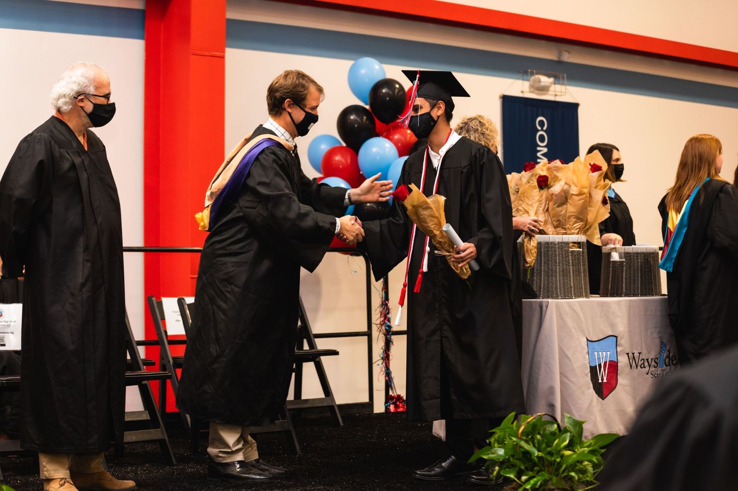 Graduate receiving diploma during commencement ceremony with red wall and decorations visible.
