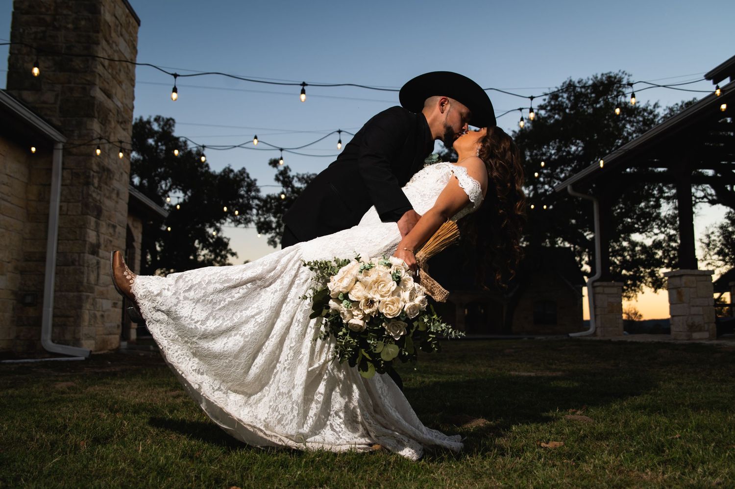 A dramatic dip pose with the bride in white dress and groom in black cowboy hat at sunset.