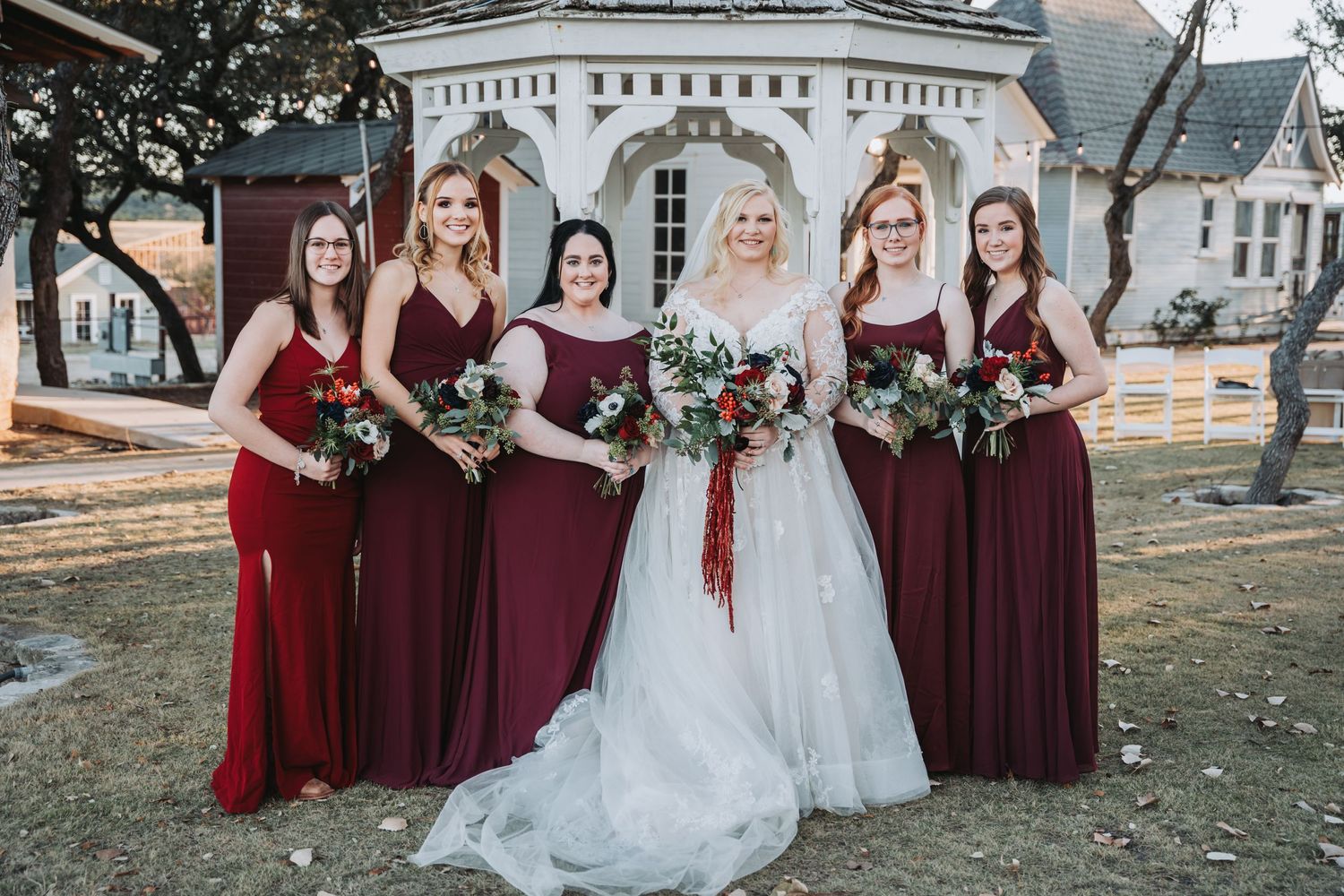 Bridesmaids in burgundy dresses pose with bouquets in front of a white gazebo.