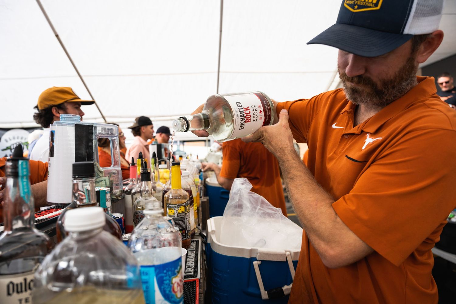 Person in orange shirt and navy cap pours drinks at outdoor bar setup.