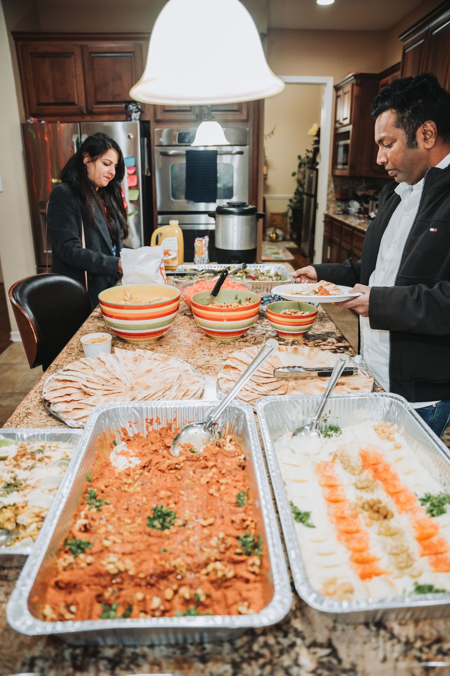 Catering buffet setup with trays of food and orange plates on kitchen counter under pendant lighting.