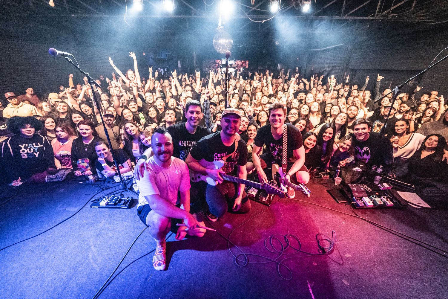 Concert crowd faces stage while performer kneels at edge with dramatic lighting effects.