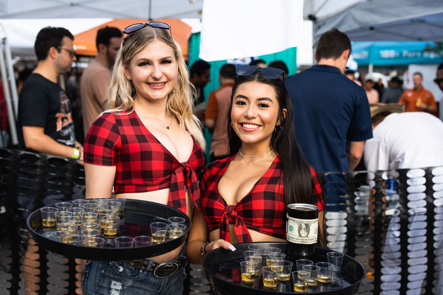 Servers in matching plaid shirts hold drink trays at an outdoor event.