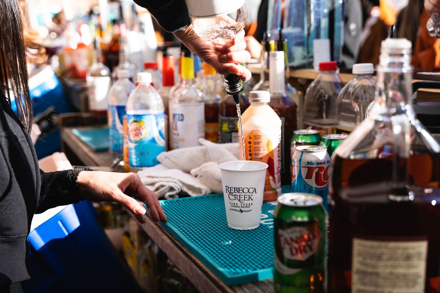 Hands preparing drinks on a bar mat surrounded by various liquor bottles and mixers.