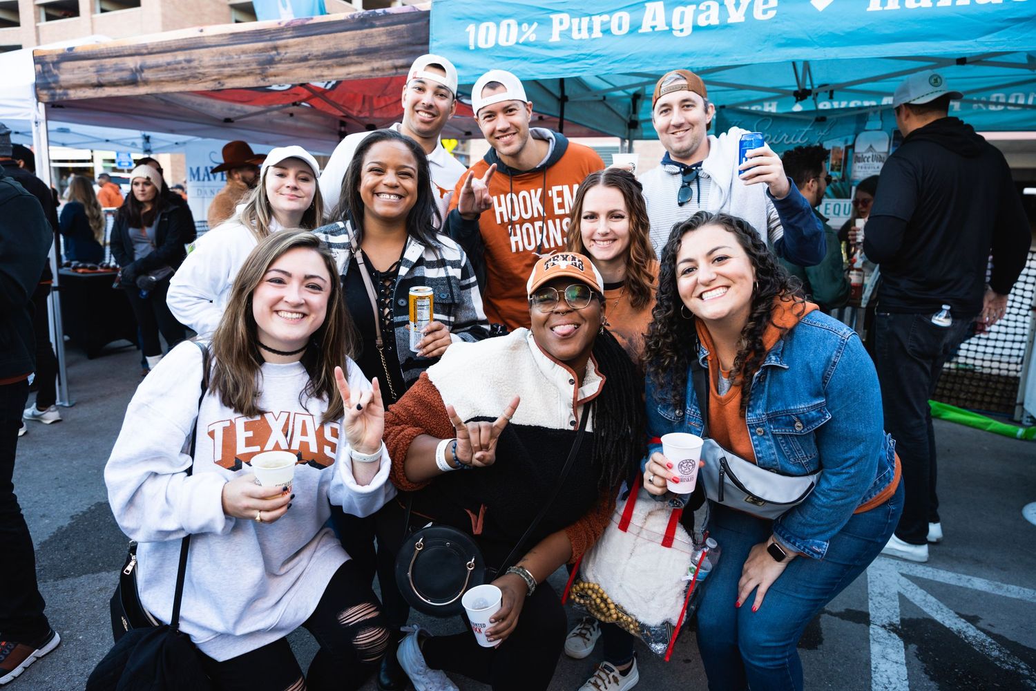 A diverse group gathers under a tent at an outdoor event, holding drinks and smiling.