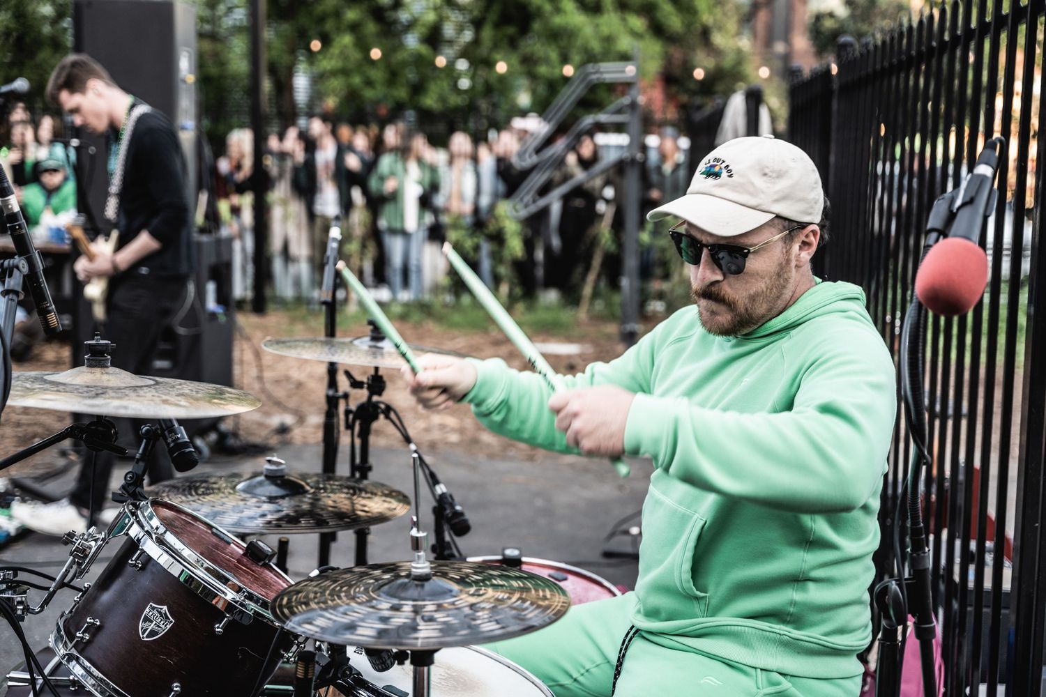 Drummer in mint green hoodie performs at outdoor concert with crowd behind iron fence.