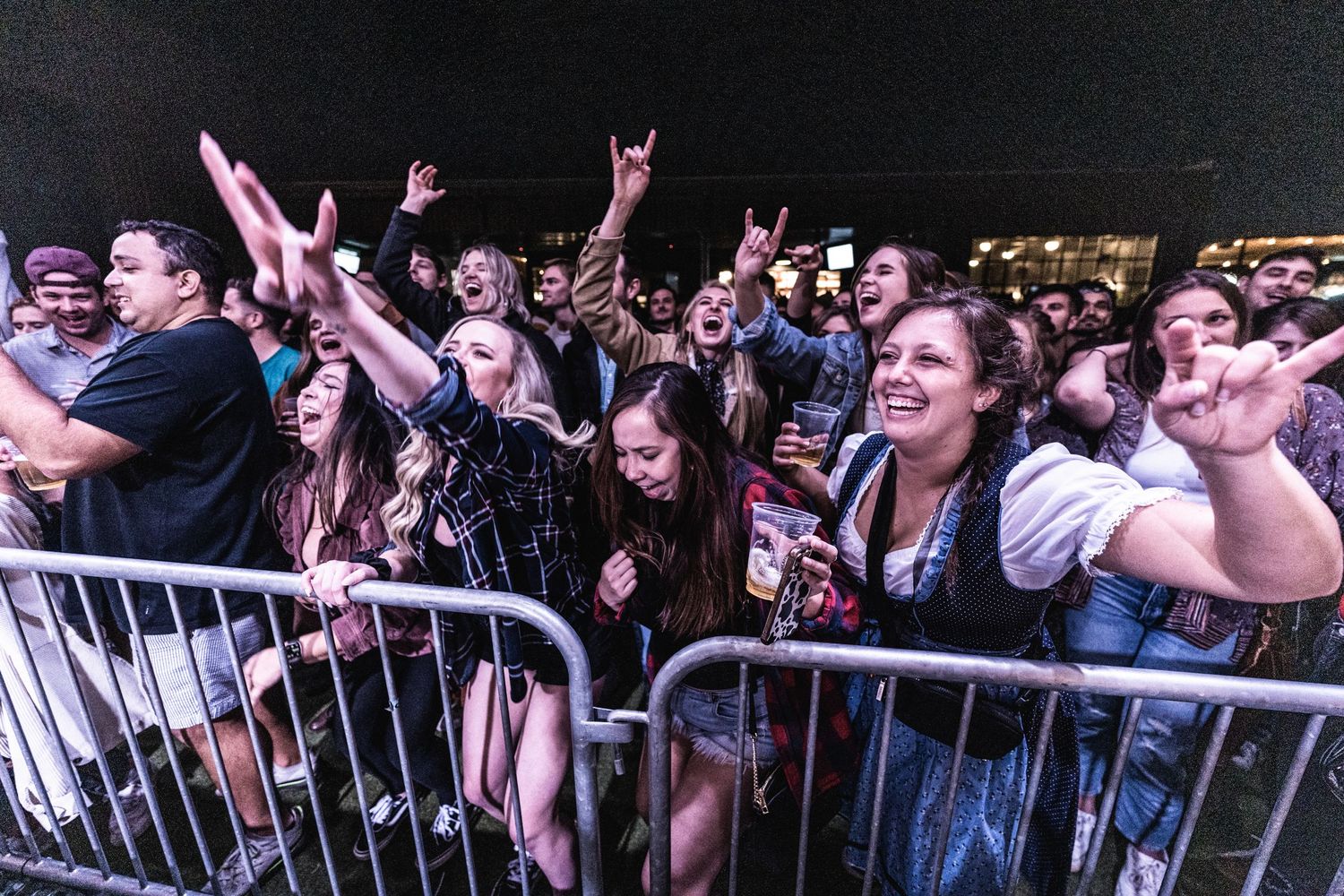 Enthusiastic crowd at outdoor concert raises hands and cheers behind metal barriers.