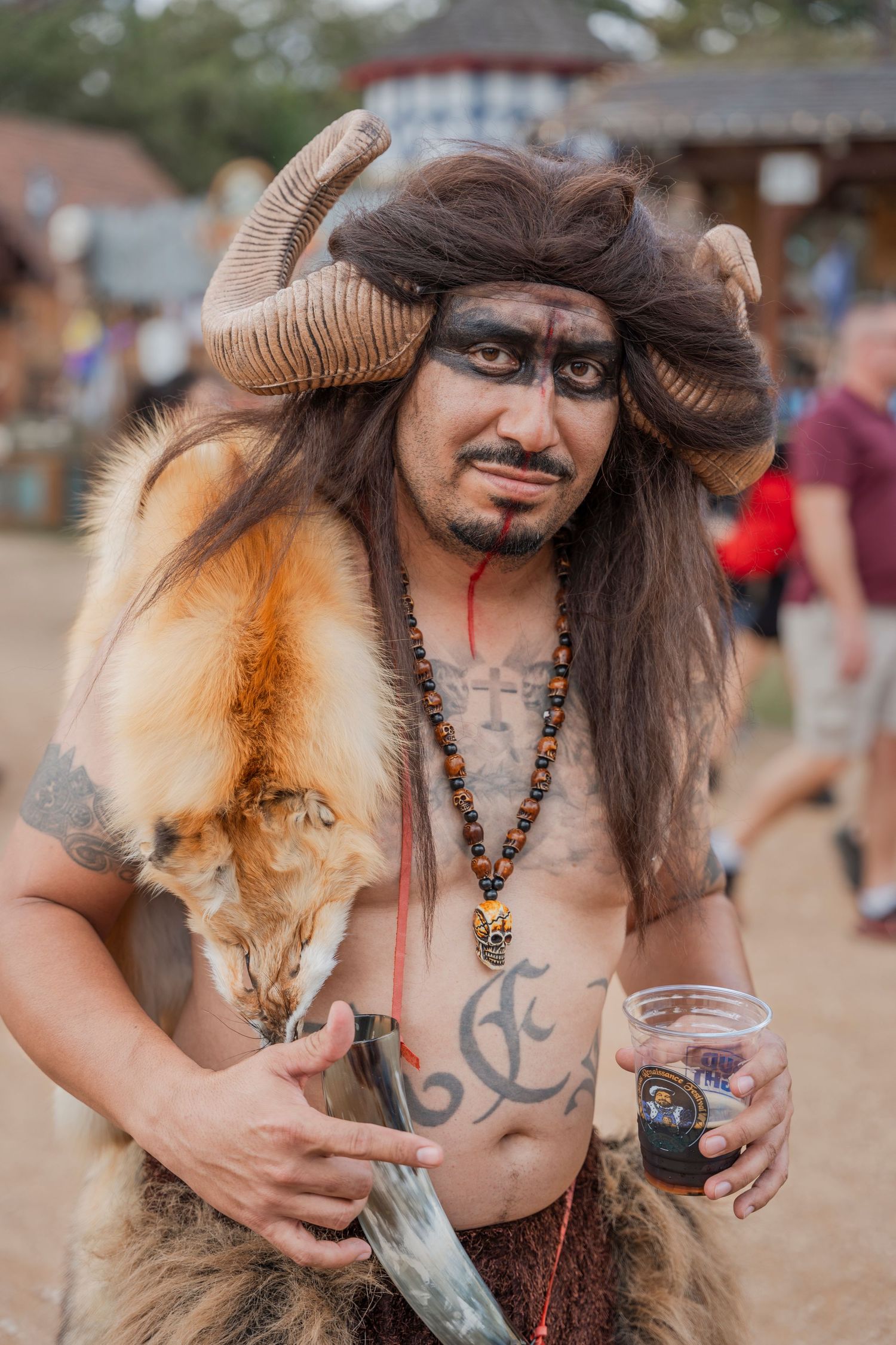 Fantasy-themed festival attendee in horned headdress and fur costume holding a drink at outdoor event.
