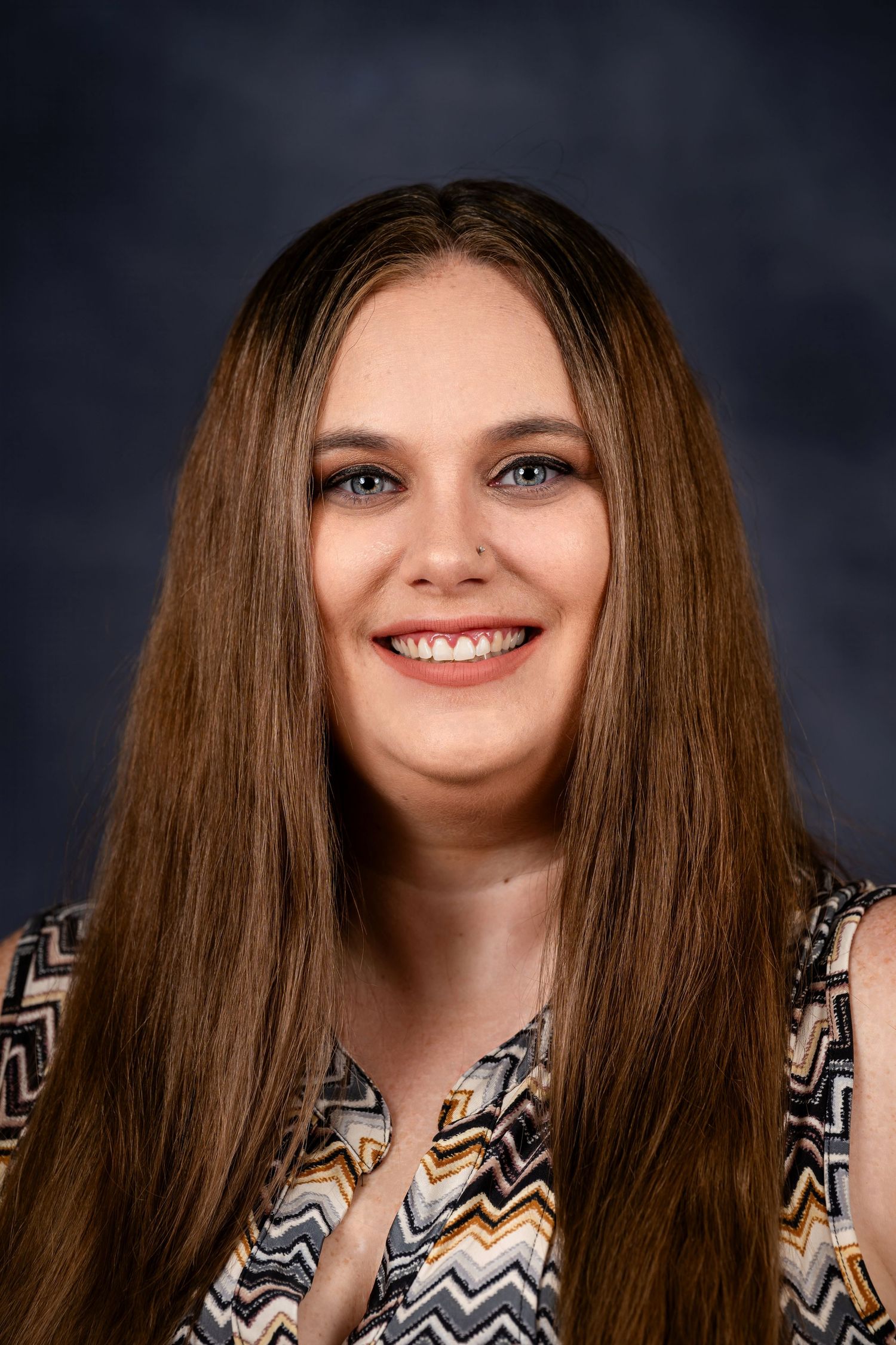 Professional headshot with long straight brown hair and patterned top against dark backdrop.