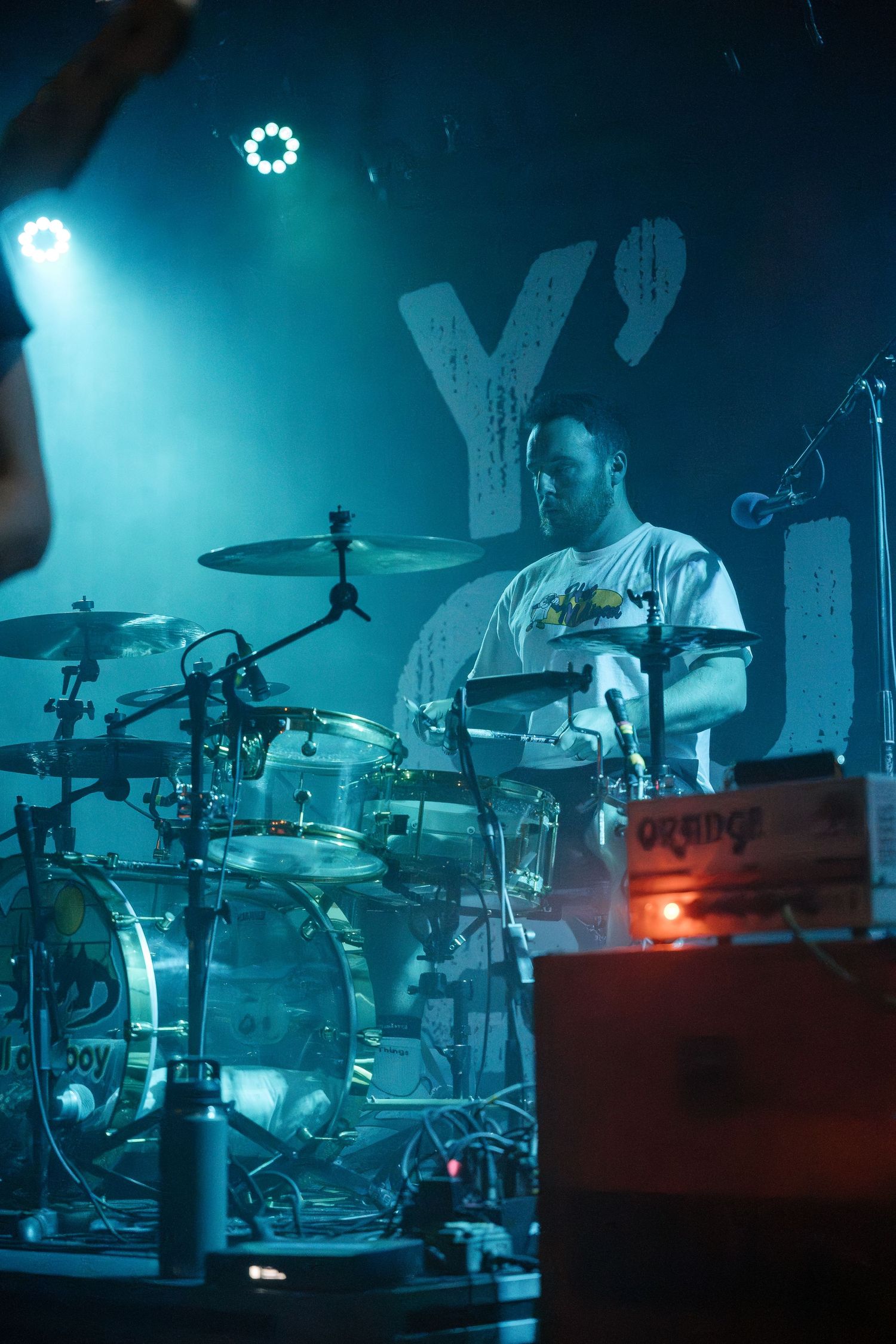 A drummer performs under blue stage lighting with a large 'Y' visible in the background.