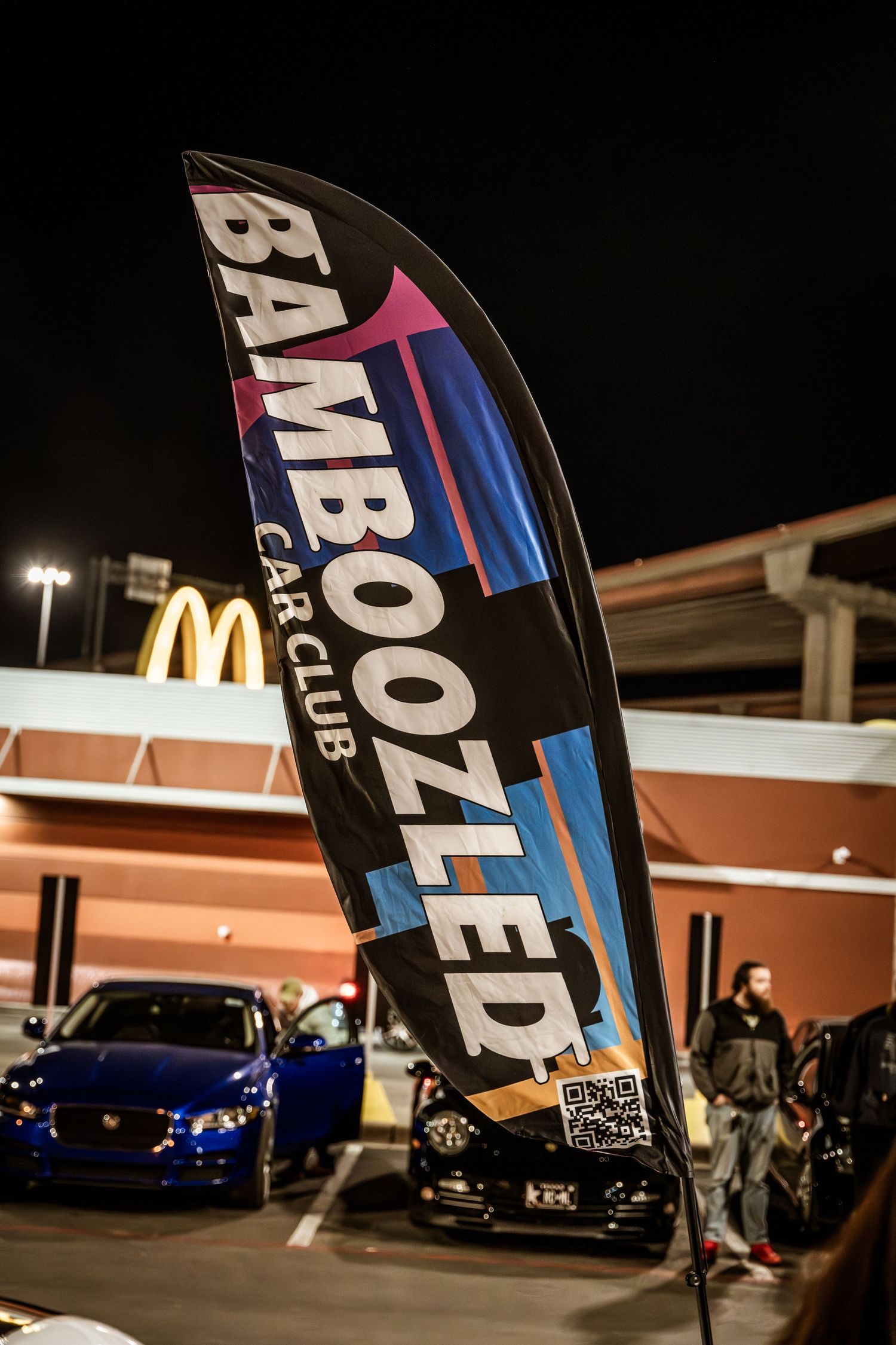 Black Bamboozled Car Club feather banner flag at night with McDonald's sign visible.