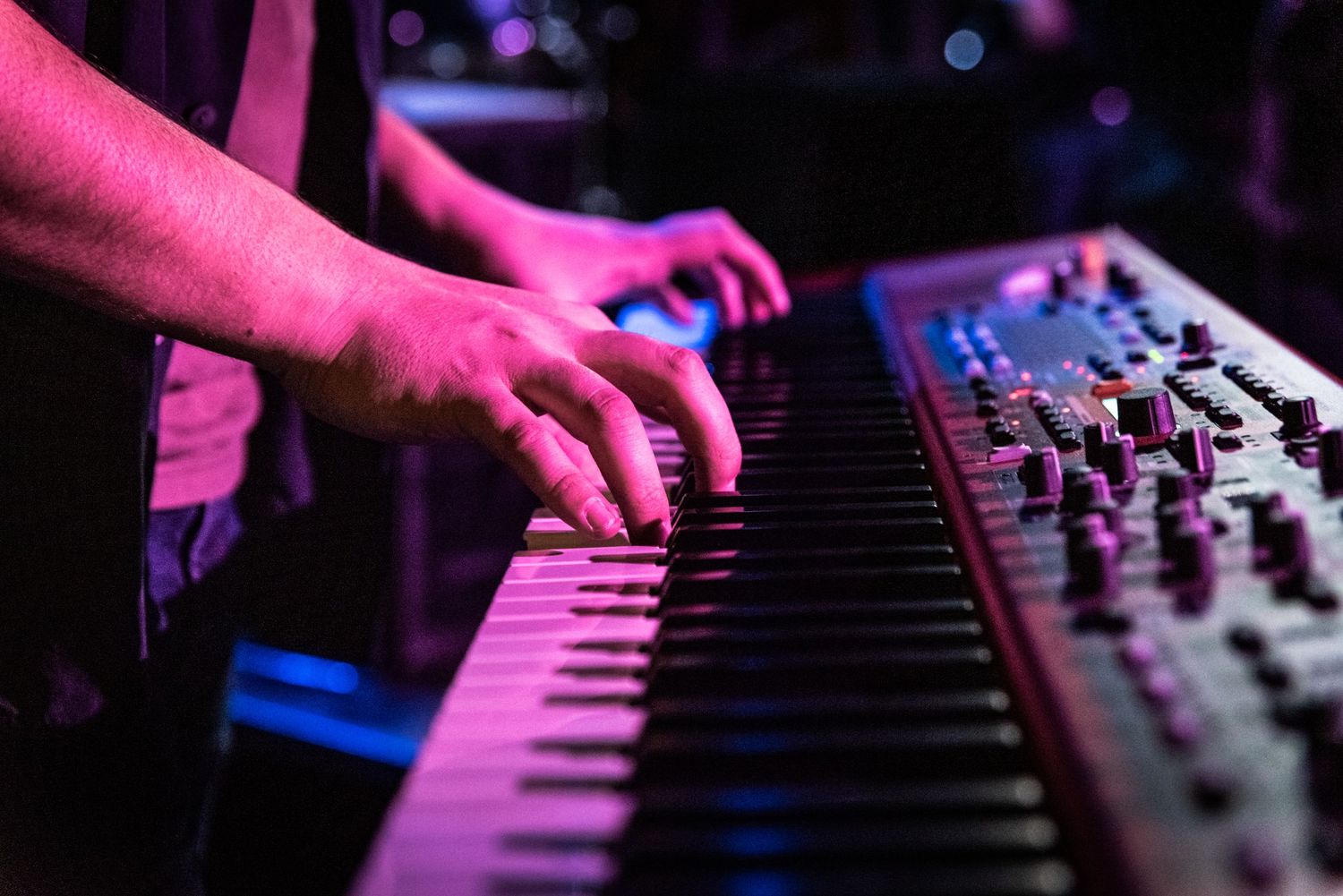 Hands playing a synthesizer keyboard with pink and blue stage lighting.