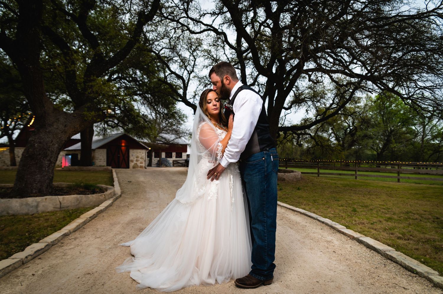 A romantic wedding couple embraces on a pathway with trees and a red building in the background.