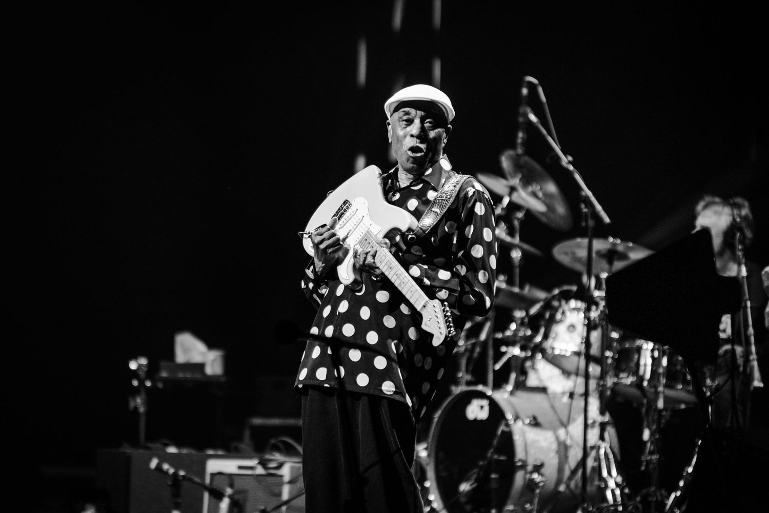 Musician in polka dot shirt performing with guitar on darkened stage with spotlights.