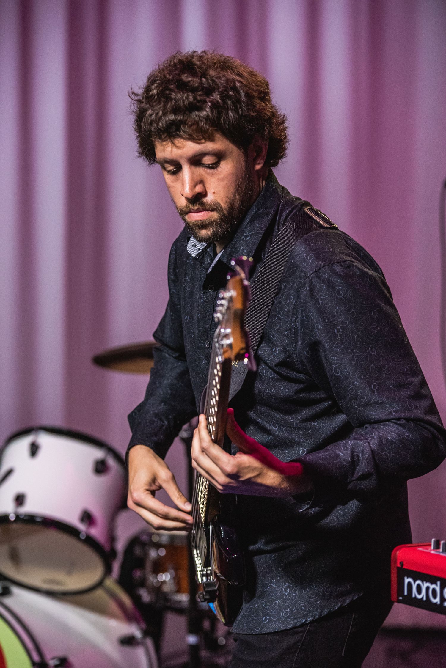 Musician plays electric guitar in dark attire against purple curtain backdrop during live performance.