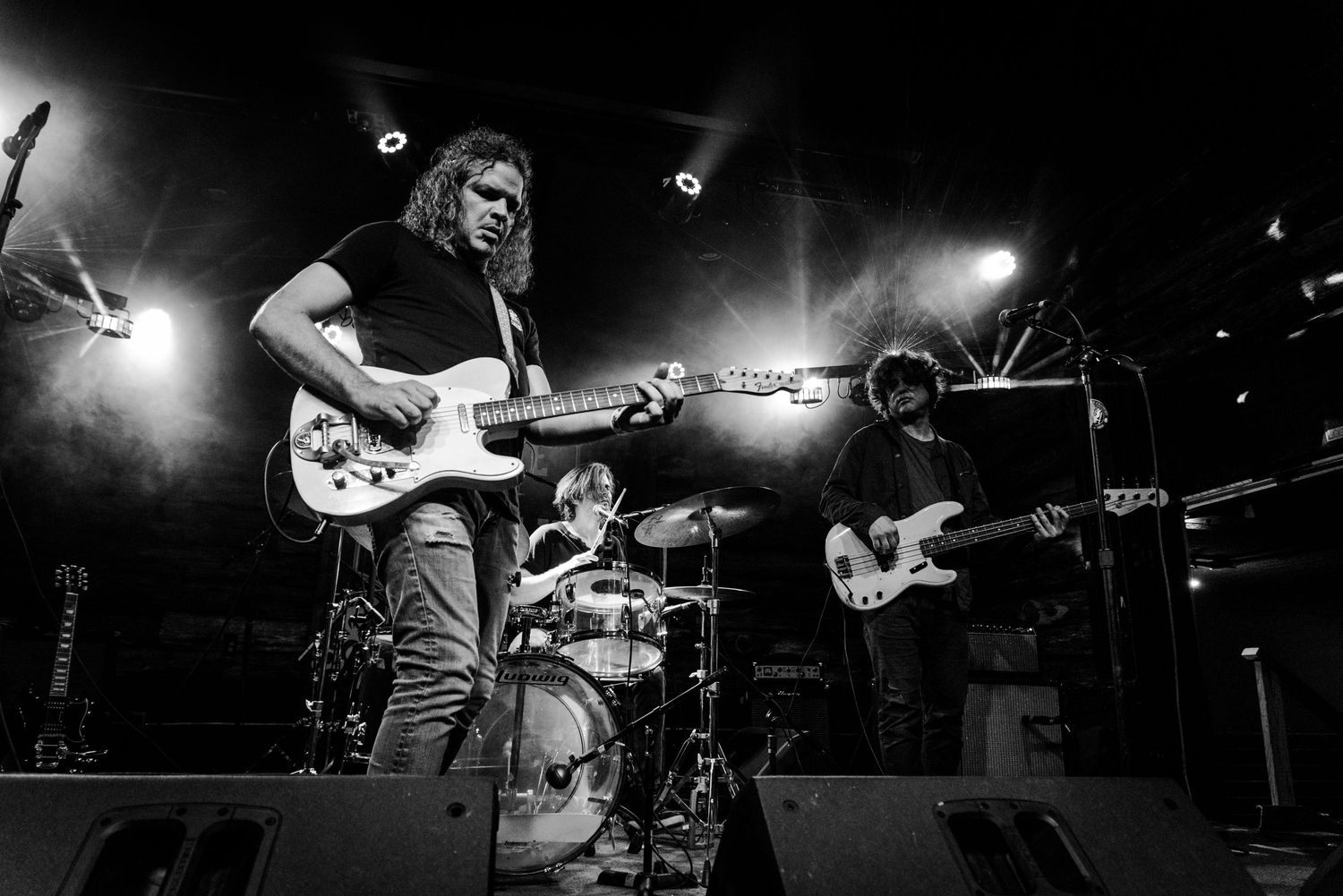 Black and white wide shot of rock band performing on stage with guitar players and drum kit.