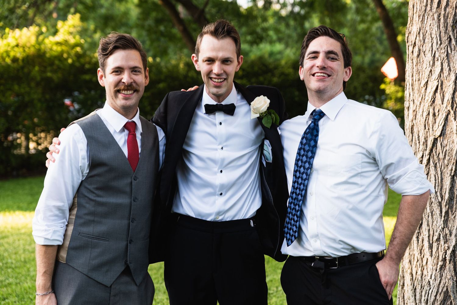 Three well-dressed men in formal attire pose together at an outdoor event.