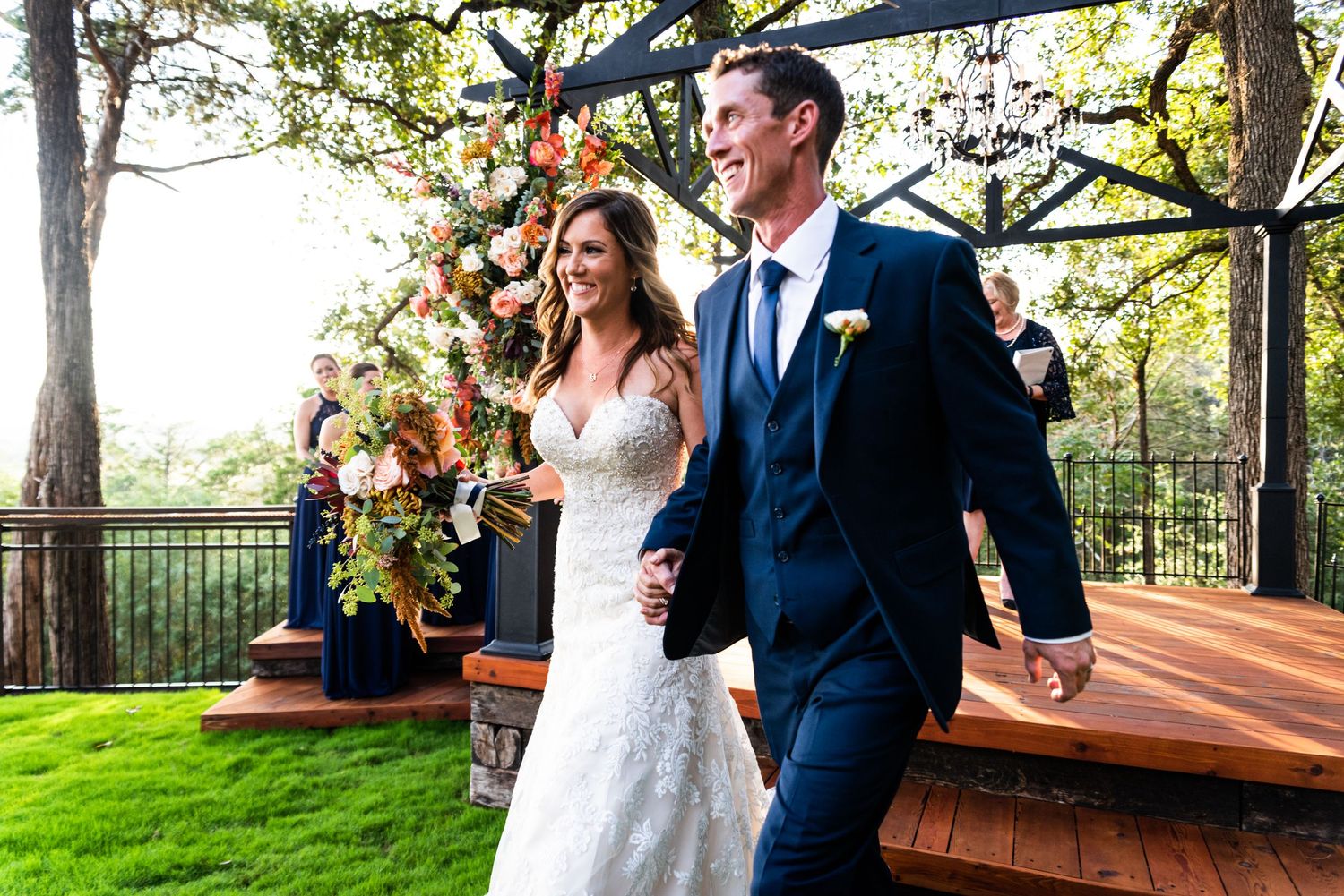 A joyful wedding couple walks across a wooden deck decorated with colorful floral arrangements.