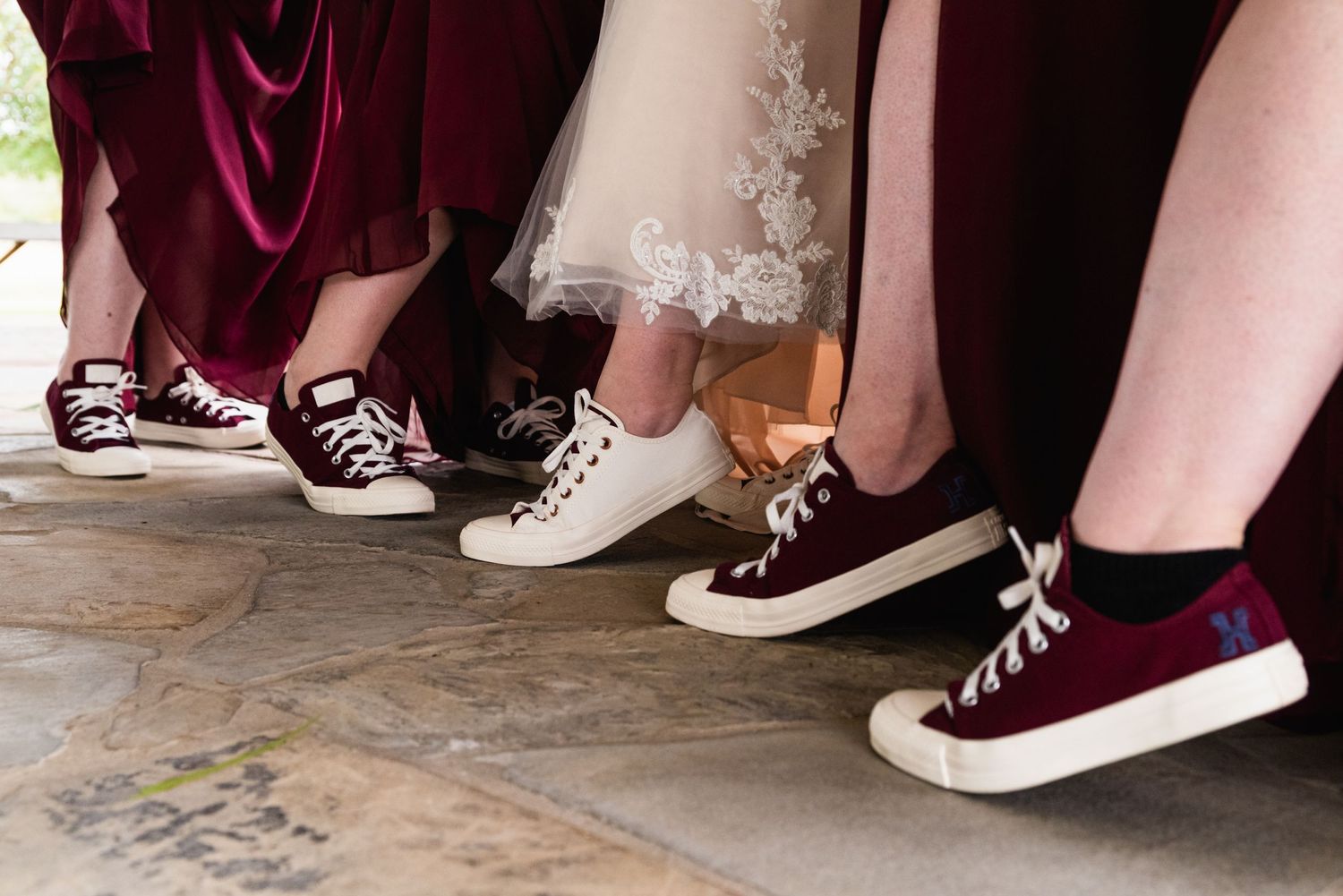 A row of feet wearing casual sneakers in burgundy and white colors against stone flooring.