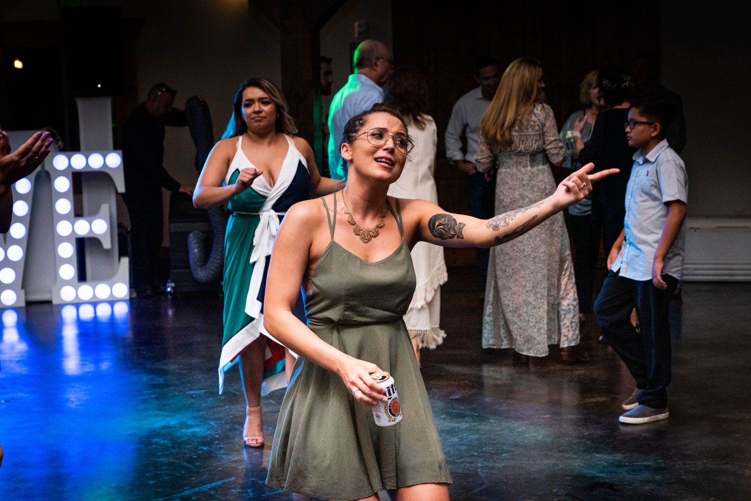 Guest in olive dress dances joyfully under blue lighting at wedding reception.