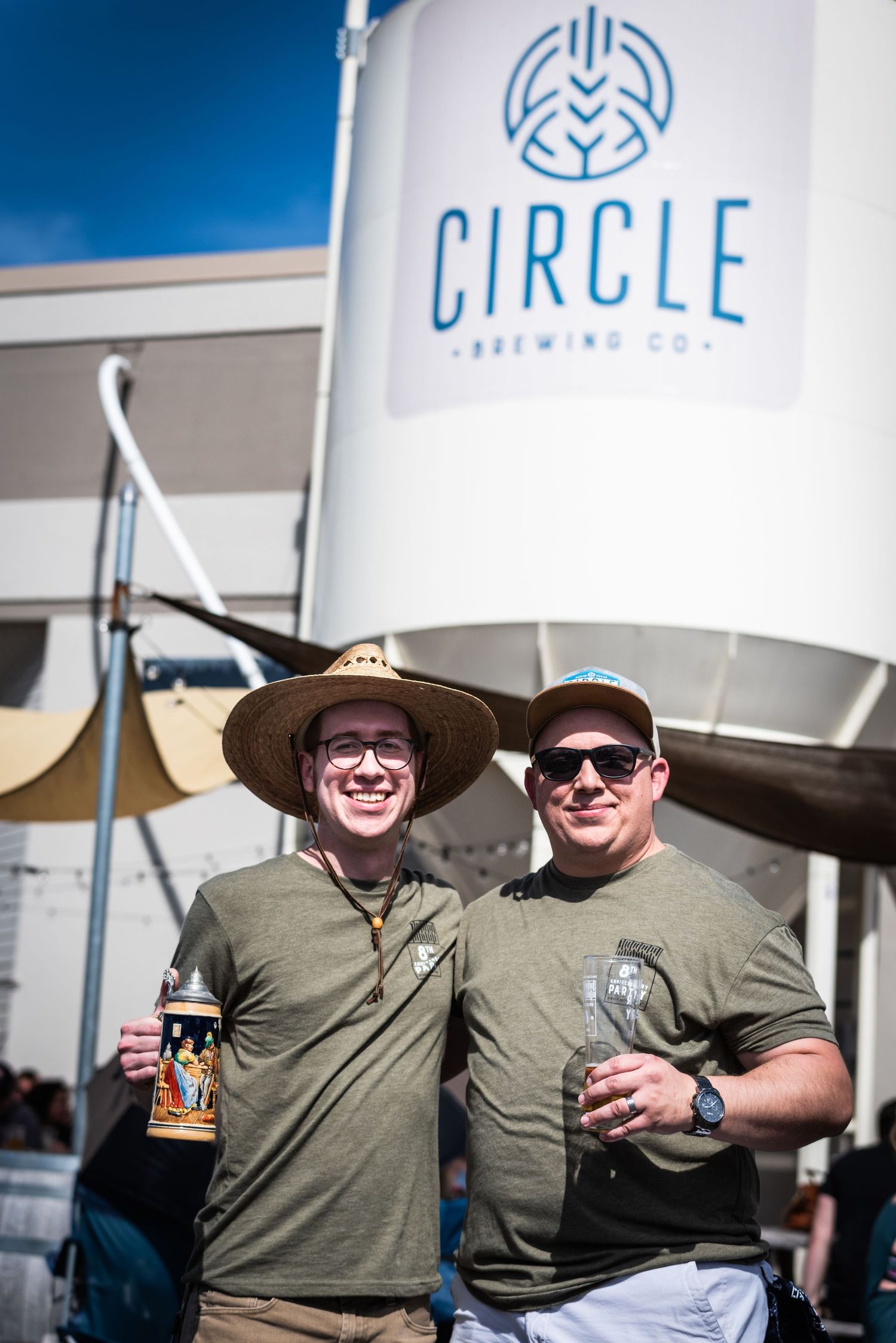 Two people in olive shirts standing in front of Circle Brewing Company logo and banner.