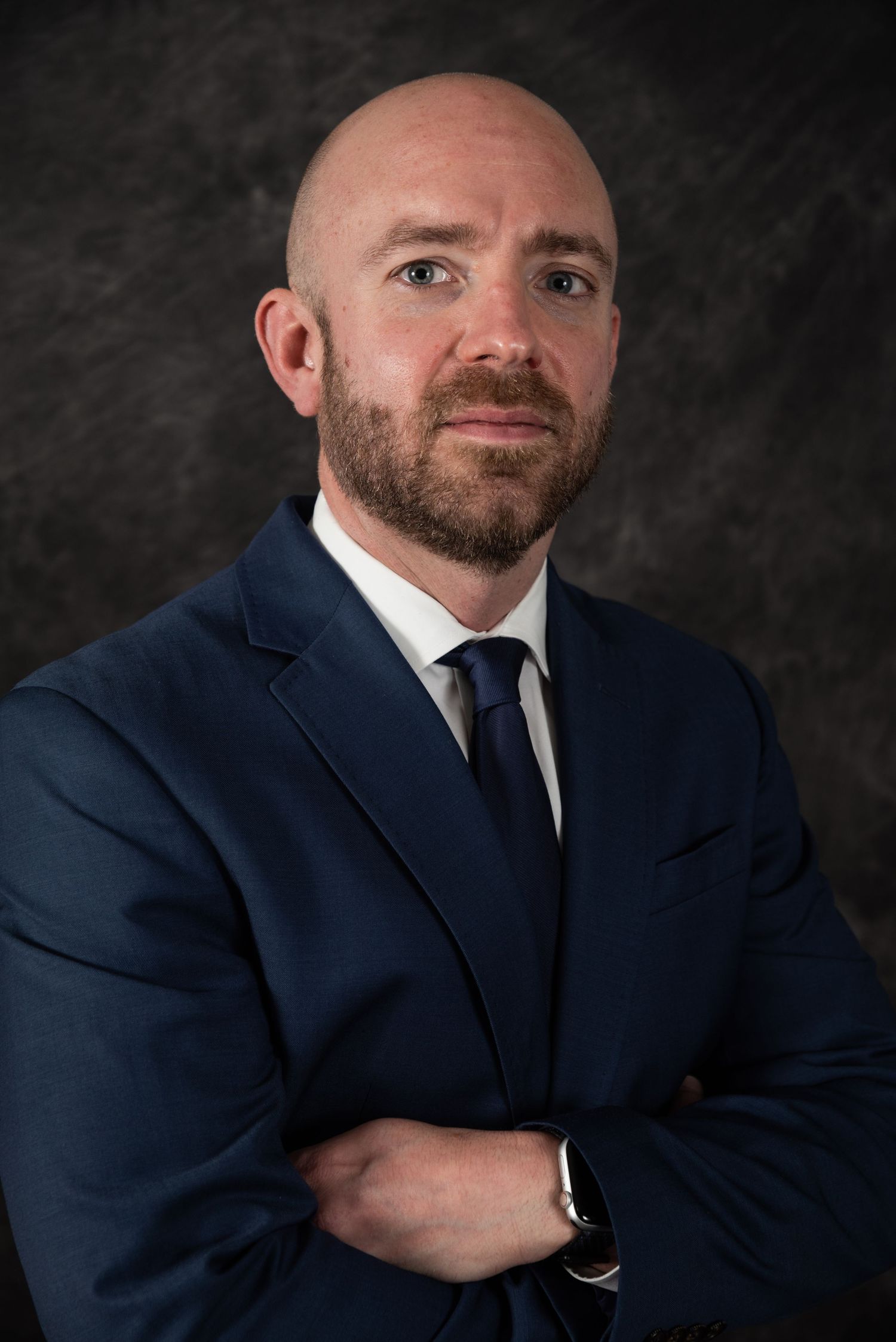 Business portrait with crossed arms pose in navy suit against dark background.