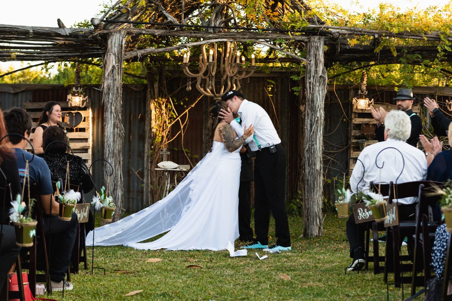 A wedding ceremony takes place under a rustic wooden pergola with guests seated on both sides.