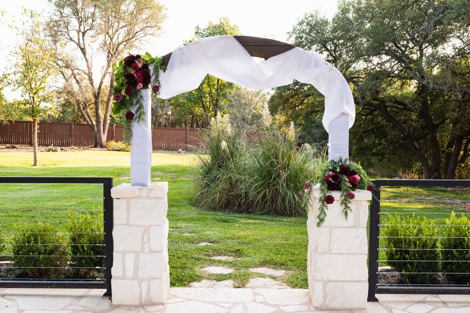 White fabric and floral wedding arch made of stone pillars in an outdoor garden setting.
