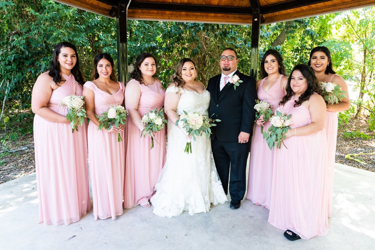 Wedding party with bridesmaids in pink dresses poses under wooden gazebo in garden setting.