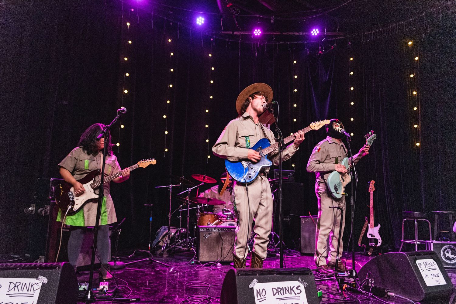 Full band performing on concert stage with purple lighting and string lights in background.
