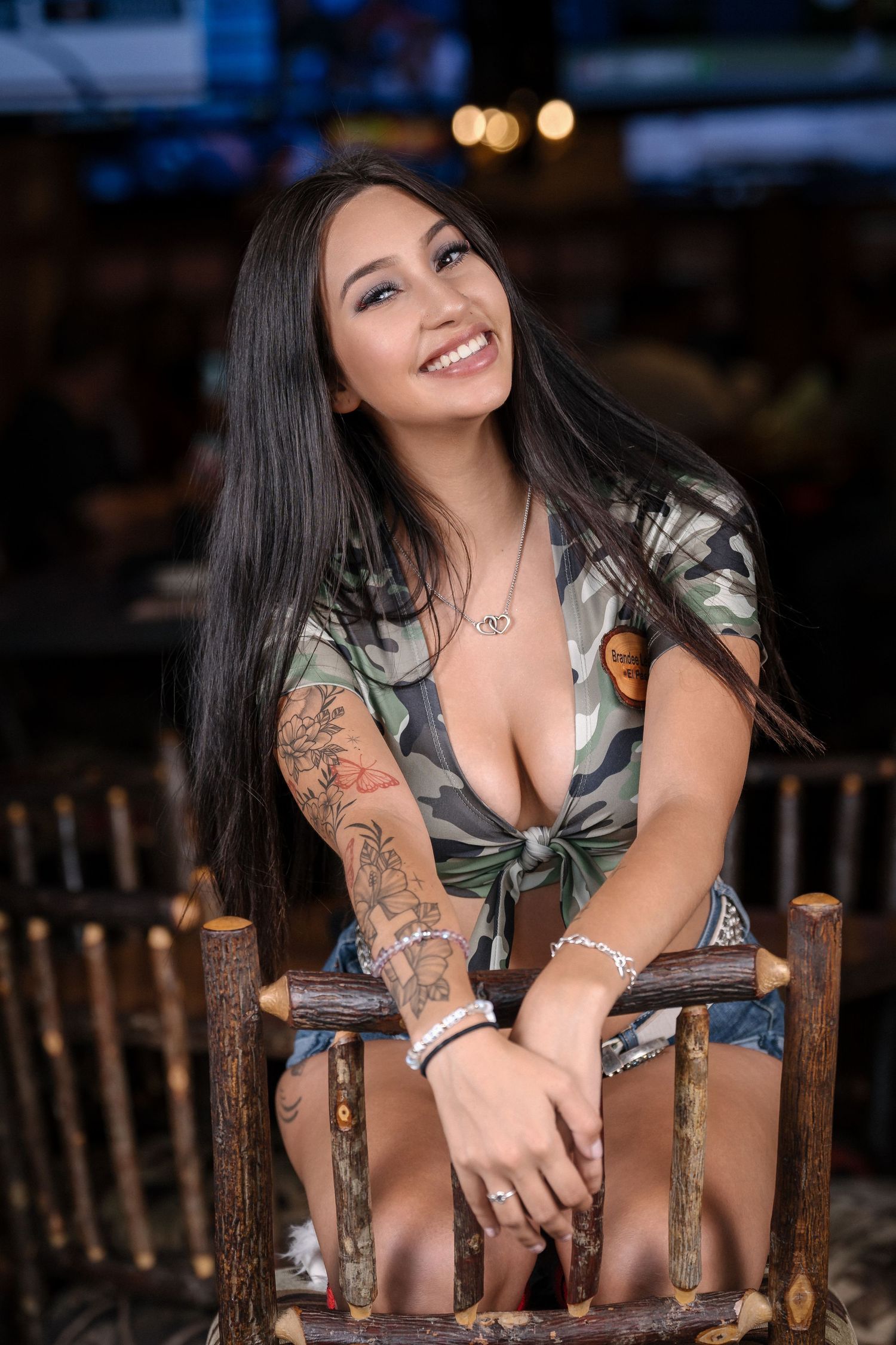 Woman in camo print top sits on wooden chair with visible tattoos and jewelry.
