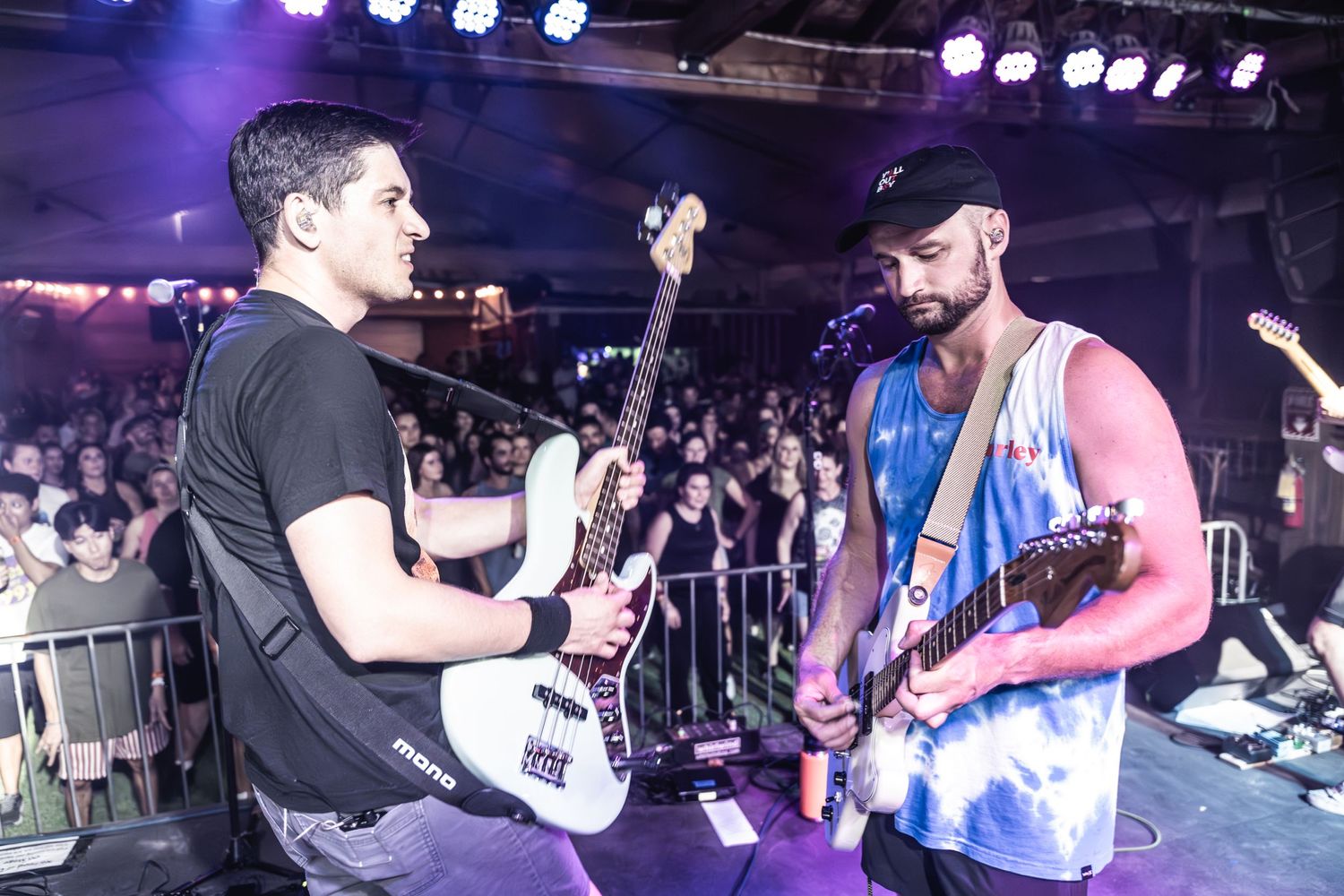 Two musicians performing with guitars on stage with crowd visible in background.