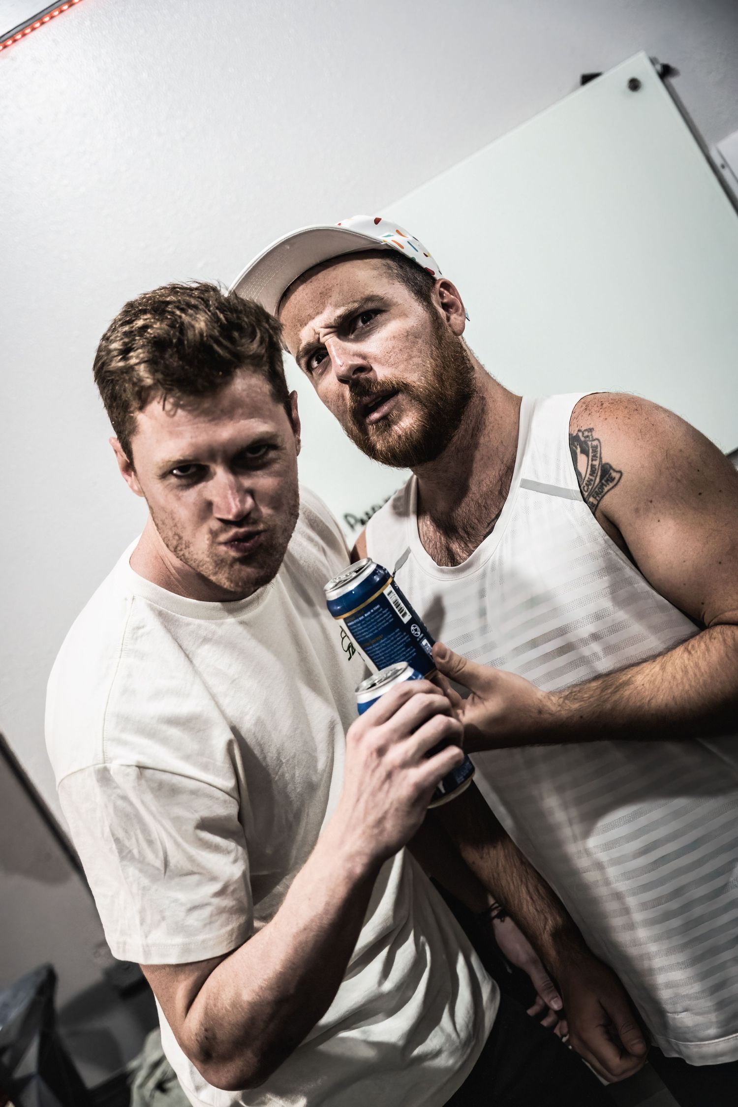 Two men in white tank tops pose together with drinks for a casual indoor photo.