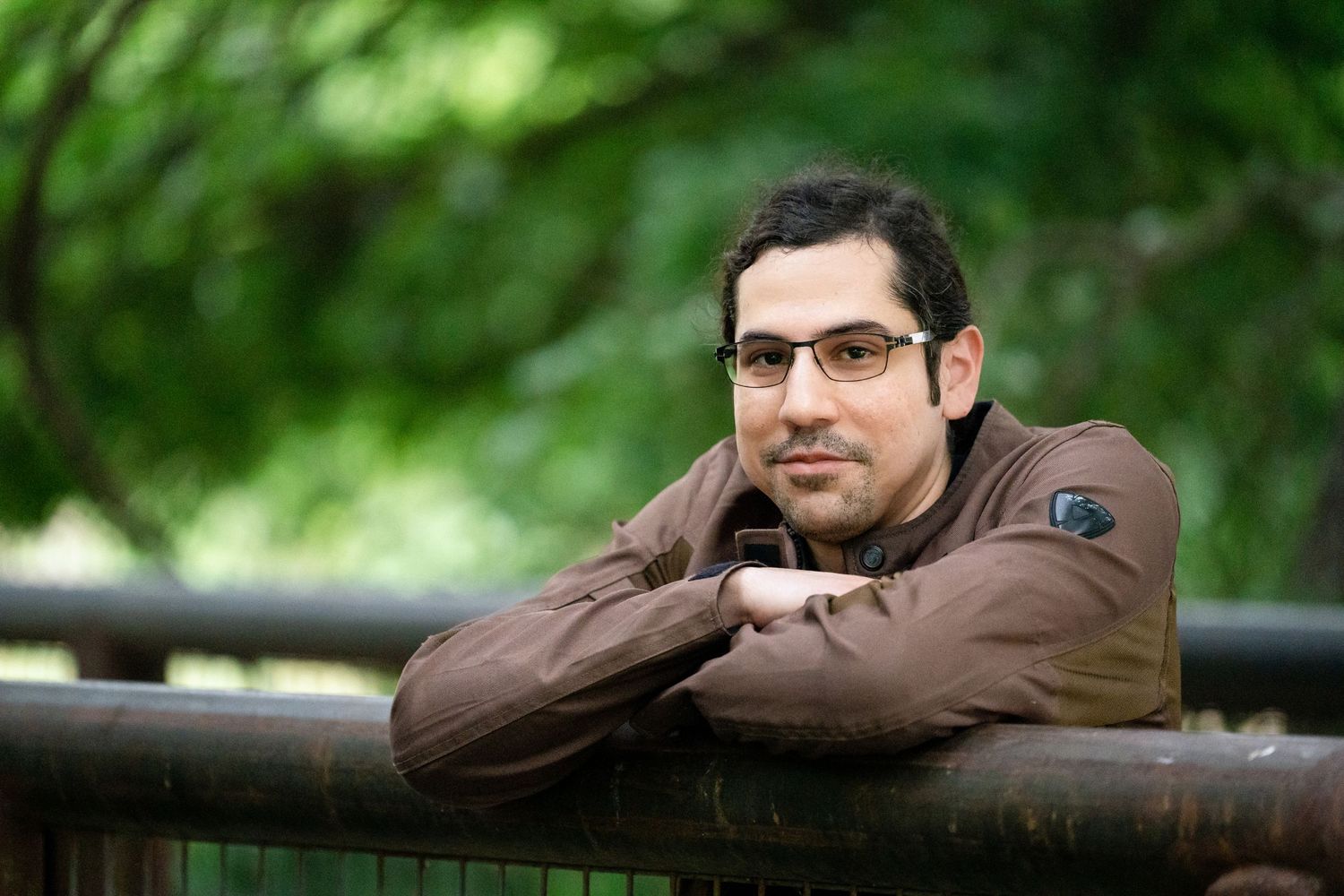 A person in glasses and brown jacket leans casually against a railing in a park setting.