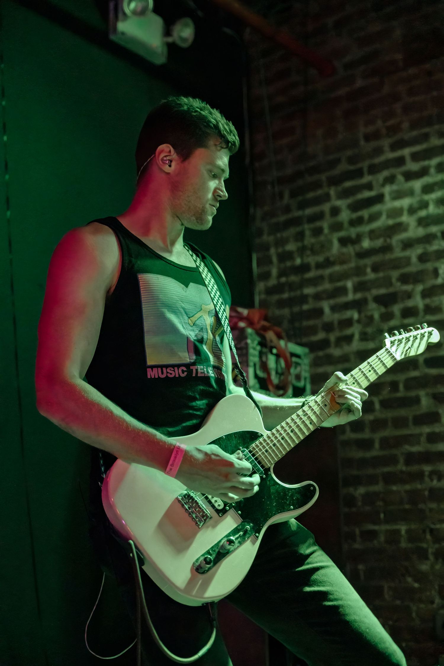 Guitarist performs with a white electric guitar under green and red stage lighting against brick wall.