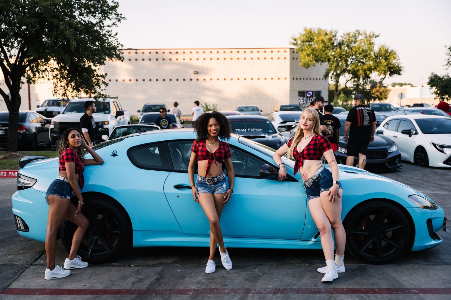 Three people in matching outfits pose against light blue sports car at sunset.