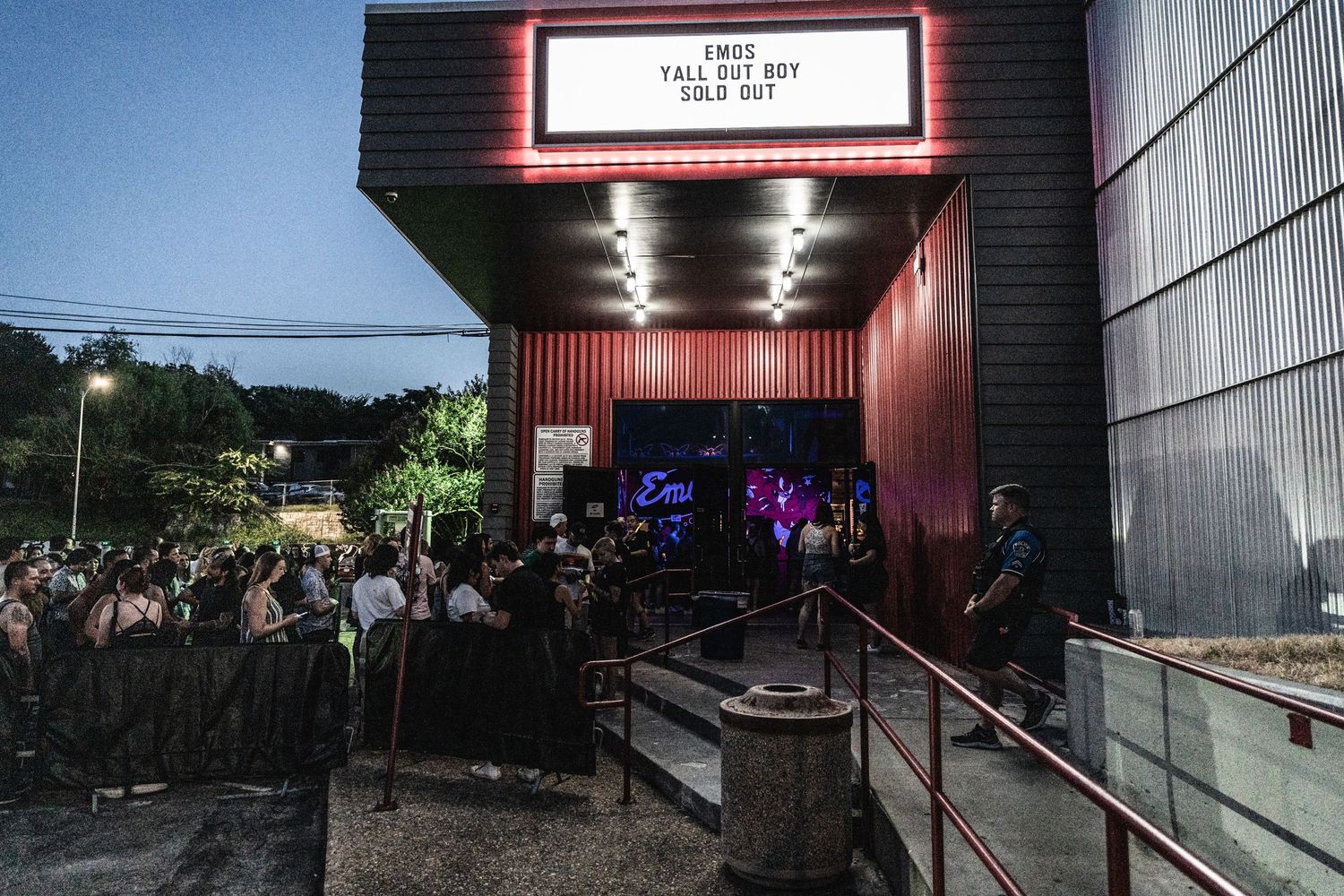 Crowd lines up outside a dimly lit venue entrance with illuminated marquee sign.