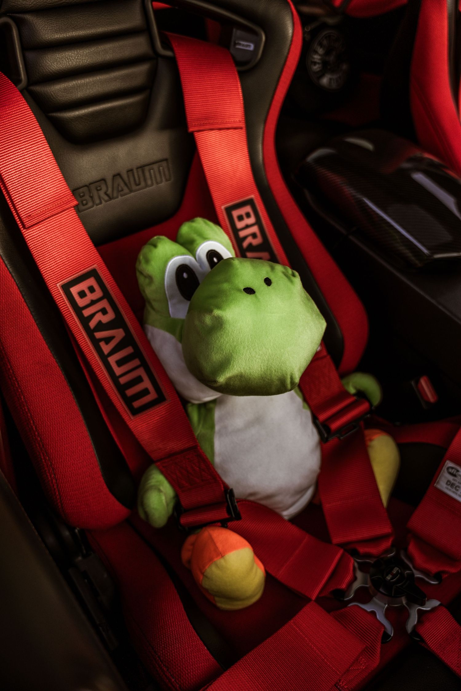 Green plush dinosaur toy sitting in a red racing seat with Braum harness straps.