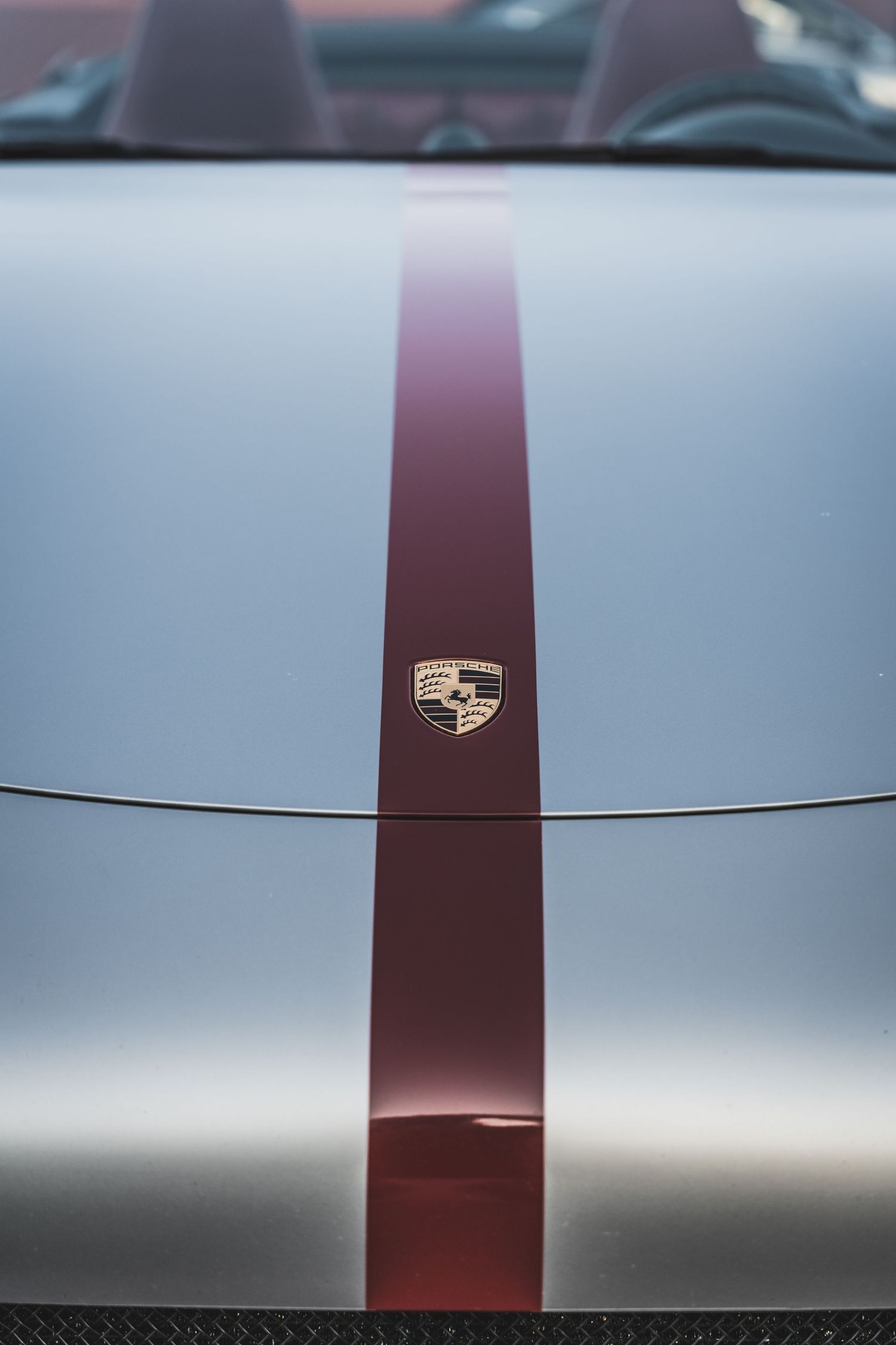 Close-up of a silver Porsche hood with burgundy racing stripe and Porsche emblem.